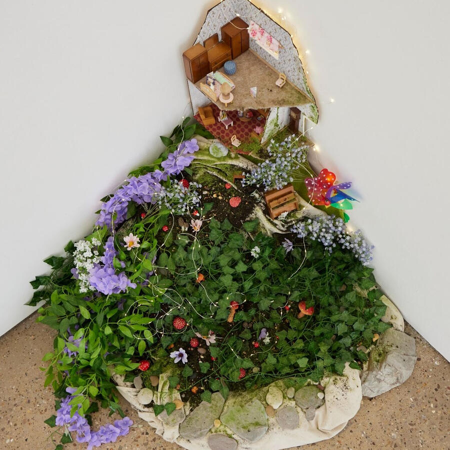 Photograph of “and she gave me magic,” an installation poem by hazyn forsythe. The piece nestles in the corner of a gallery space, resembling a cross-section of a tiny, overgrown home. A dollhouse-style interior sits against the wall, complete with miniatu