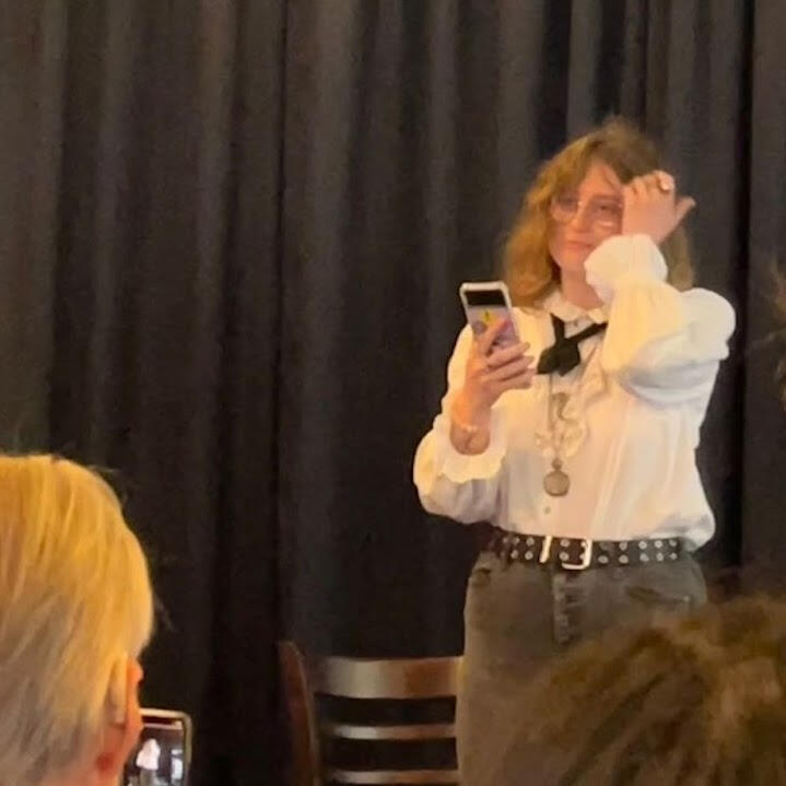 A second photo of hazyn forsythe reading their piece sealed from Monstera at a 2022 event. They are standing in front of a black curtain, holding a phone, and wearing a white ruffled shirt with a black ribbon tie, layered necklaces, and studded belt. Audie