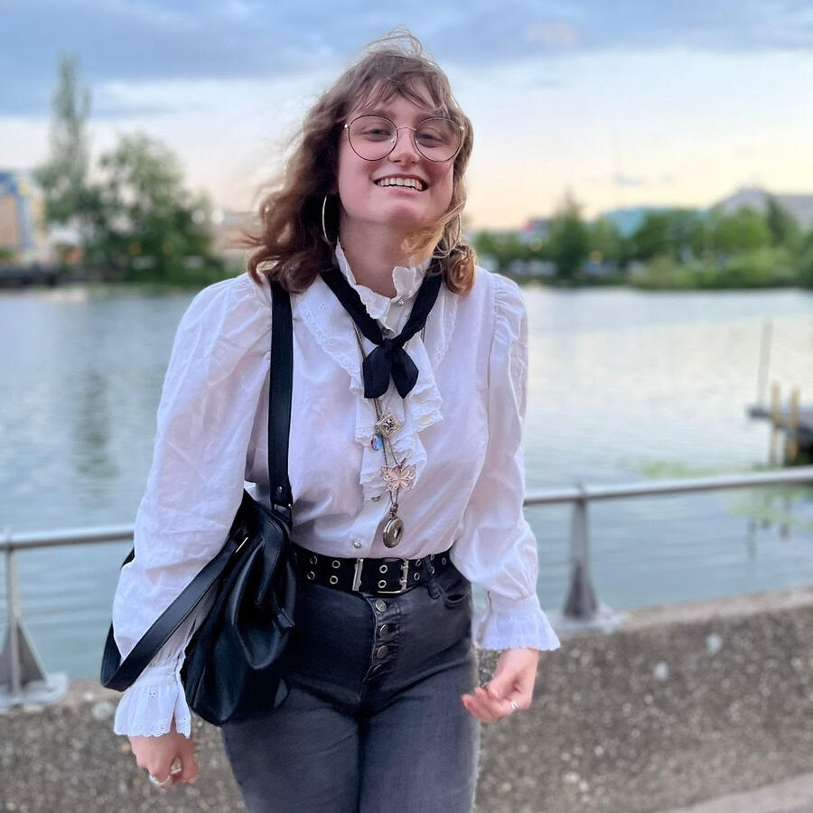 hazyn forsythe smiling proudly after their 2022 reading, standing by the water at Lincoln’s Brayford. They wear a white ruffled shirt with layered necklaces and a black ribbon tie, high-waisted black jeans, and round glasses. Their hair is tousled by the w