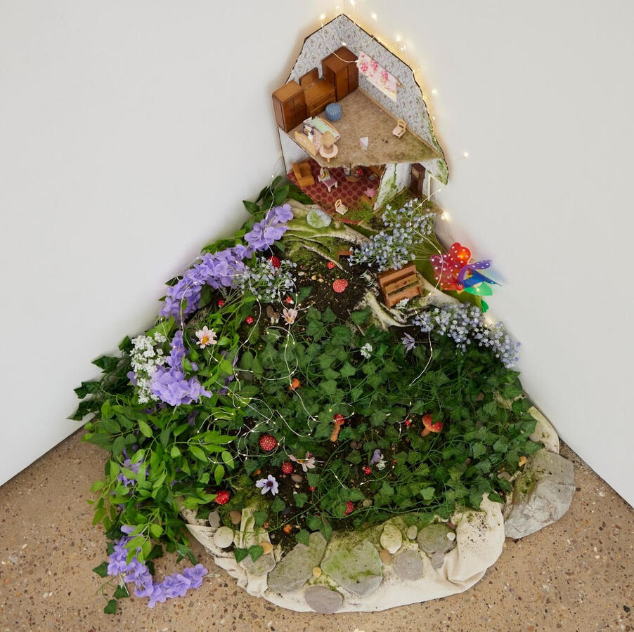 Photograph of “and she gave me magic,” an installation poem by hazyn forsythe. The piece nestles in the corner of a gallery space, resembling a cross-section of a tiny, overgrown home. A dollhouse-style interior sits against the wall, complete with miniatu