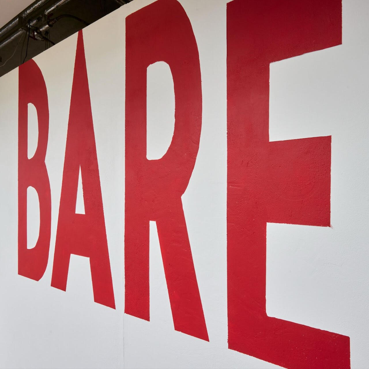 Exhibition title wall for BARE (2023), where and she gave me magic by hazyn forsythe was displayed. The word “BARE” is painted in large, bold red capital letters across a white wall, extending toward the viewer in a dynamic perspective.